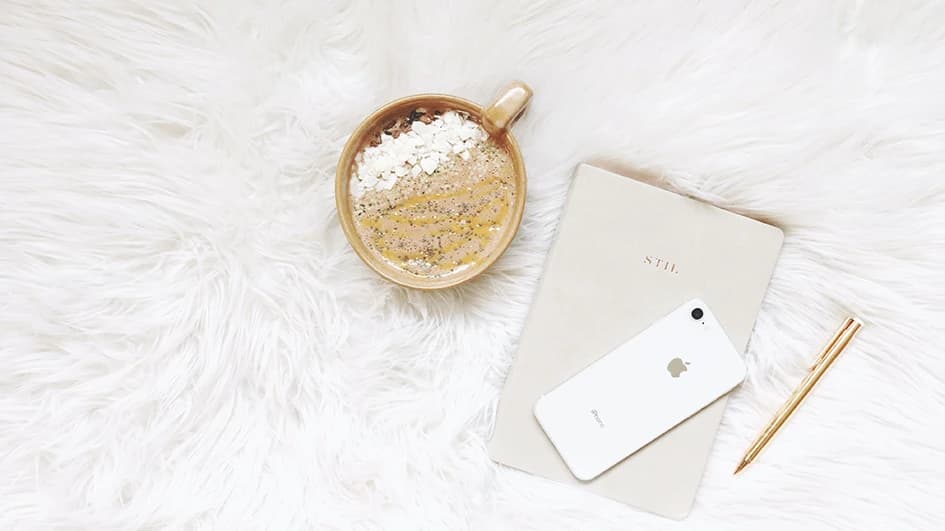Tasse de latte, carnet beige « Stil », iPhone blanc et stylo doré posés sur une fourrure blanche