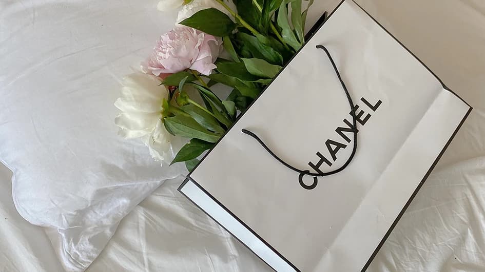 Sac shopping blanc Chanel posé sur des draps blancs avec un bouquet de pivoines roses et blanches