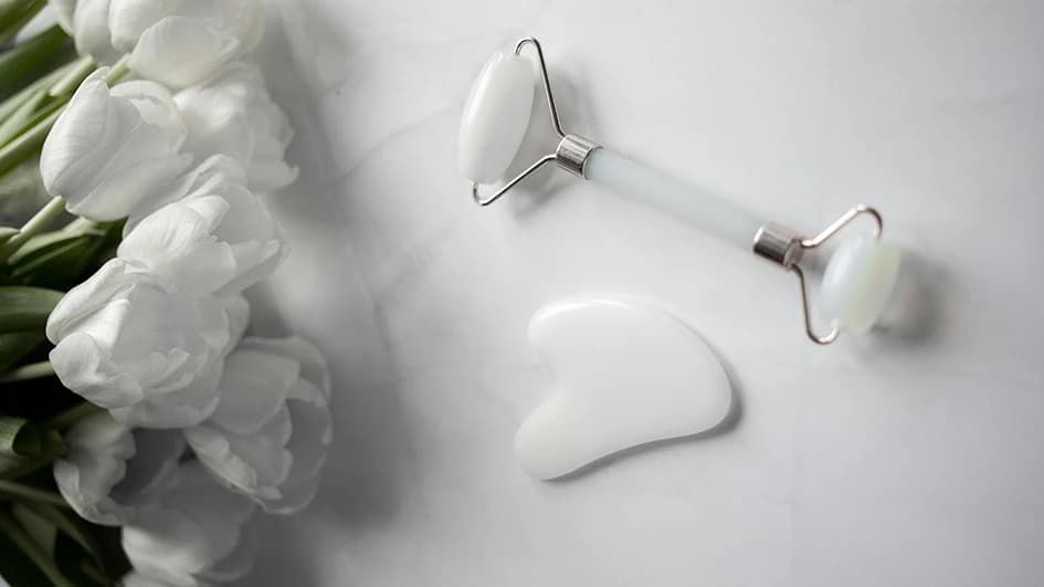 Rouleau de massage et pierre gua sha en jade blanc sur du marbre, à côté d'un bouquet de tulipes blanches