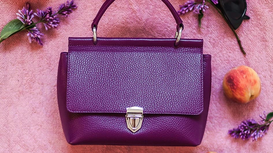 Sac à main en cuir violet prune posé sur un tissu rose, entouré de brins de lilas et d'une pêche