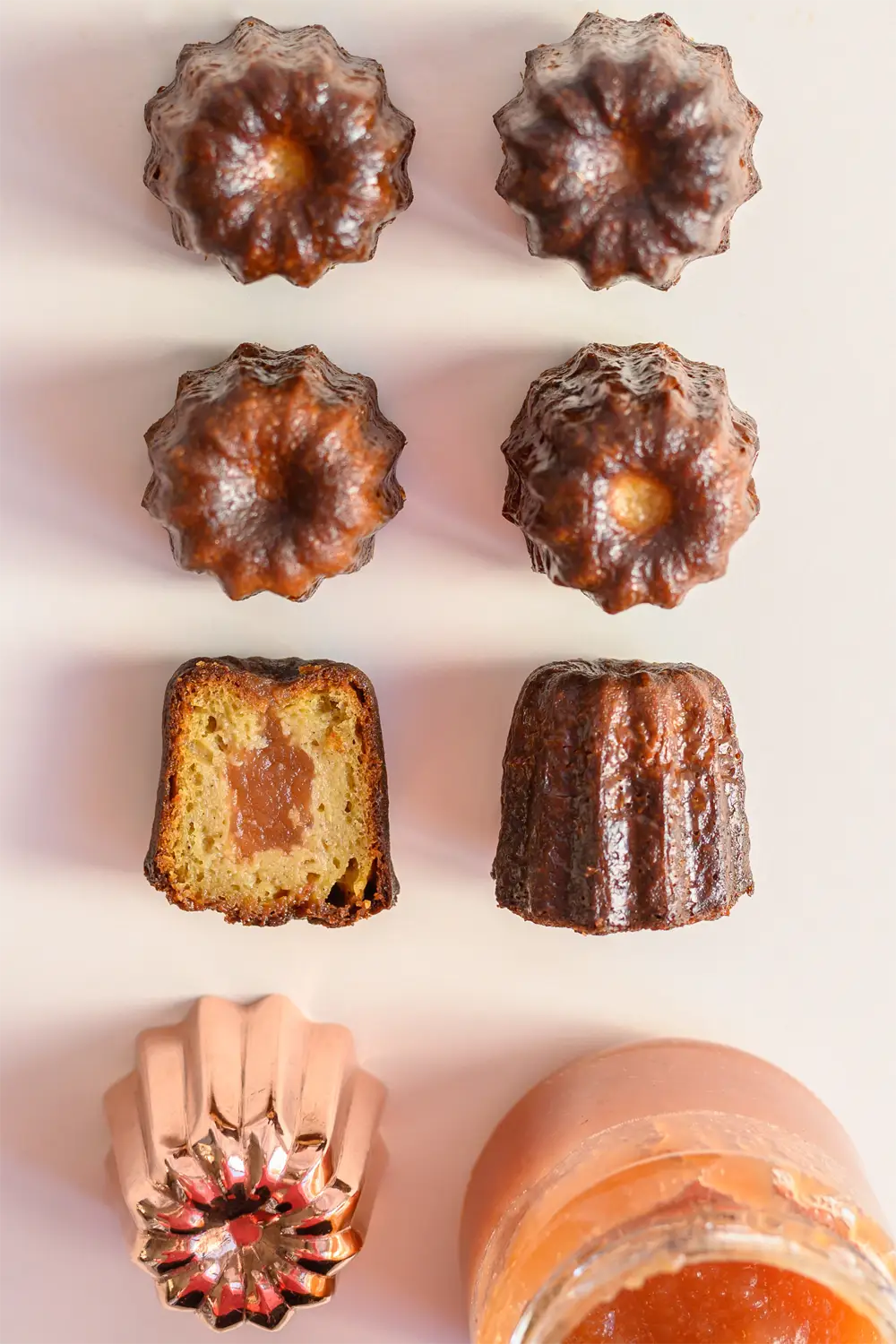 Étude de cas : Six cannelés des Gourmandises d’Énora vus de dessus et en coupe, moule en cuivre et pot de confiture sur fond rose pâle