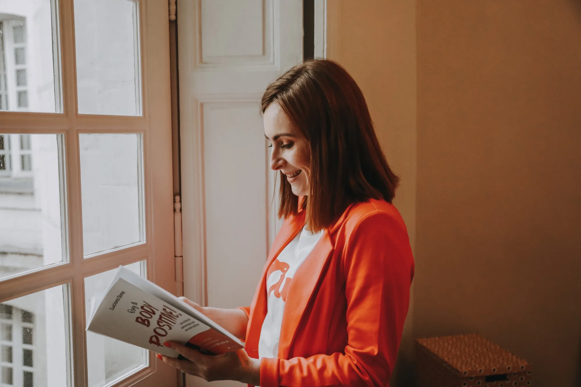 Fanny Métivier, graphiste à Nantes spécialisée en design éditorial, feuillette un livre imprimé près d’une fenêtre, en veste orange